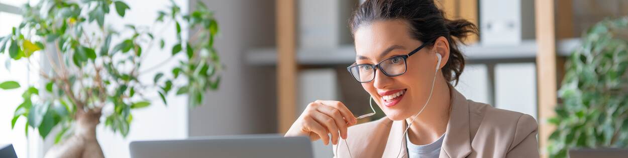 Vrouw met bril zit lachend achter haar laptop met oordopjes in die verbonden zijn aan haar laptop