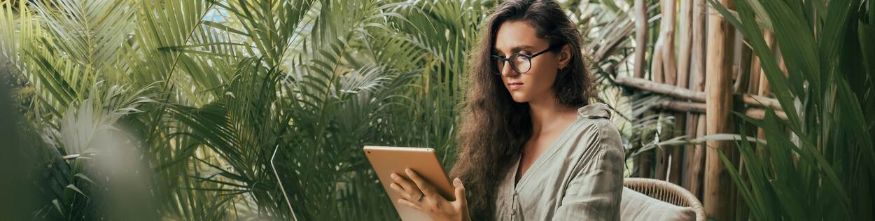 Vrouw met krullend haar en een bril is omgeven door planten en werkt op haar laptop